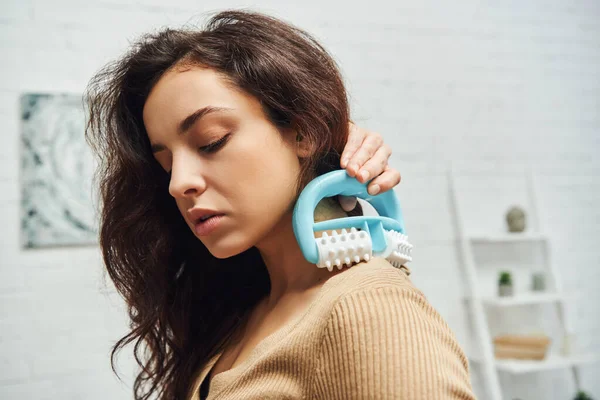 Portrait of young brunette woman in casual jumper holding handled massager and doing shoulder self-massage in blurred house, enhancing self-awareness and body relaxation concept, balancing energy — Stock Photo