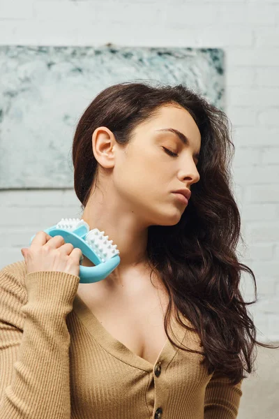 Young brunette woman with closed eye holding handled massager while doing self-massage of lymphatic nodes in blurred house, enhancing self-awareness and body relaxation concept, balancing energy — Stock Photo