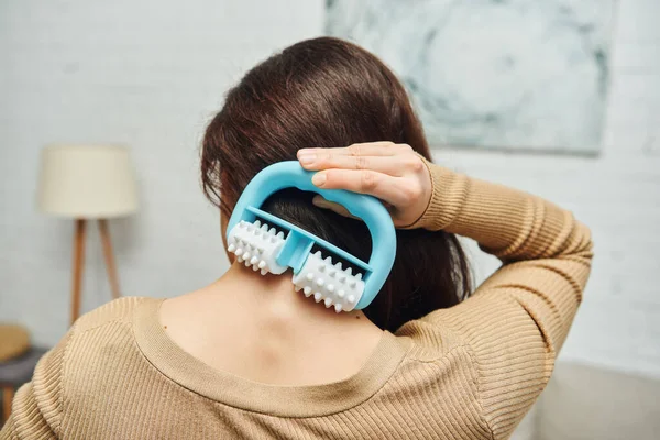 Back view of brunette woman massaging neck and back with modern handled massager while standing in blurred living room, lymphatic system support and home-based massage, tension relief — Stock Photo