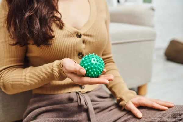Tension relief, cropped view of blurred brunette woman in casual clothes holding manual massage ball while sitting near couch at home, lymphatic system support and home-based massage — Stock Photo