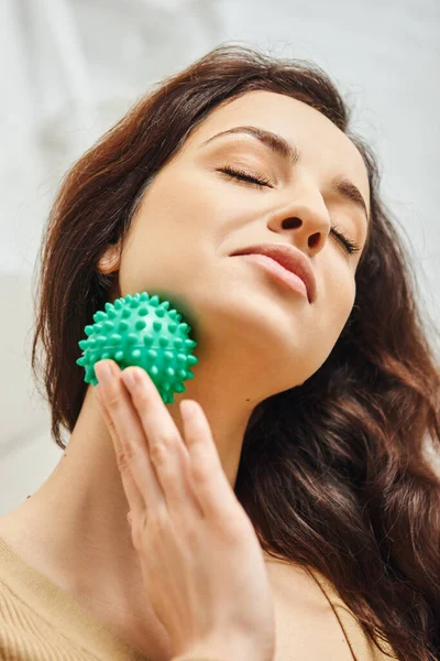 Portrait de jeune femme brune heureuse aux yeux fermés massant le cou avec boule de massage manuelle à la maison, soutien du système lymphatique et massage à domicile, énergie équilibrante — Photo de stock
