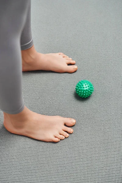 Ansicht des manuellen Massageballs in der Nähe einer barfüßigen Frau, die zu Hause auf einer Fitnessmatte steht, Körperentspannung und ganzheitliche Wellnesspraktiken, die Energie ausgleichen — Stockfoto
