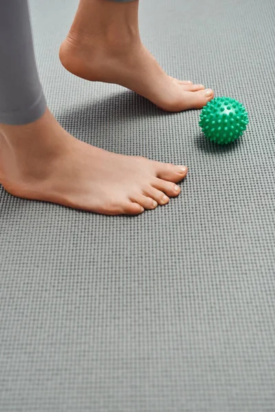 Vista ritagliata della donna scalza in piedi sul tappeto fitness vicino alla palla da massaggio manuale a casa, rilassamento del corpo e pratiche olistiche di benessere, energia riequilibrante — Foto stock