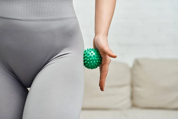 Cropped view of young woman in sportswear massaging hip with manual massage ball in blurred living room, body relaxation and holistic wellness practices, tension relief — Stock Photo