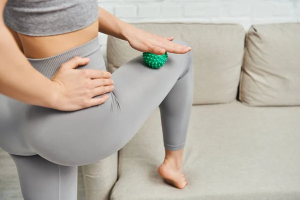 Cropped view of young woman in sportswear massaging leg with manual massage ball near couch in living room, holistic wellness practices and body relaxation concept, tension relief — Stock Photo