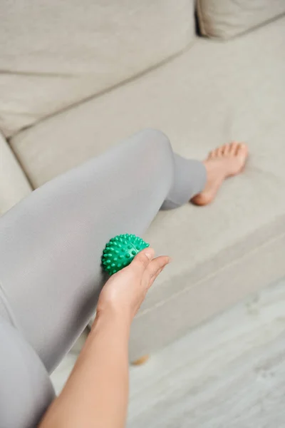 Visão superior da mulher em sportswear massagem perna com bola de massagem manual perto de sofá embaçado na sala de estar, práticas de bem-estar holístico e conceito de relaxamento corporal, alívio da tensão — Fotografia de Stock