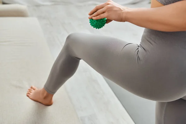 Vista cortada da jovem mulher em sportswear segurando bola de massagem manual e de pé perto do sofá na sala de estar em casa, práticas holísticas de bem-estar e conceito de relaxamento corporal, alívio da tensão — Fotografia de Stock