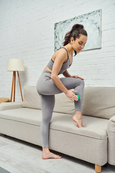 Young brunette woman in sportswear massaging leg with manual massage ball while standing near couch at home, holistic wellness practices and body relaxation concept, tension relief — Stock Photo