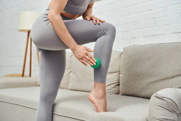 Cropped view of barefoot young woman in sportswear massaging leg with manual massage ball while standing near couch at home, holistic wellness practices and body relaxation concept, tension relief — Stock Photo