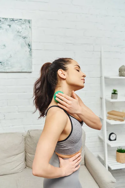 Young brunette woman in sportswear massaging neck with manual massage ball and standing in living room, balancing energy and holistic healing concept, myofascial release — Stock Photo