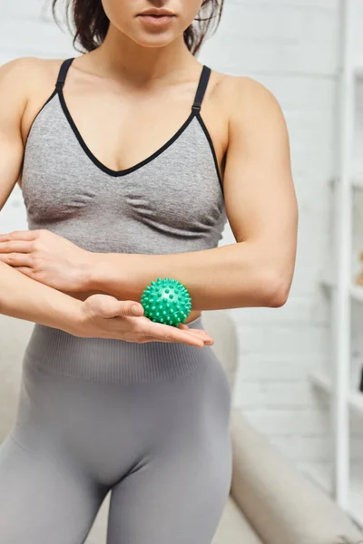 Cropped view of young woman in sportswear holding manual massage ball and standing in blurred living room, balancing energy and holistic healing concept, myofascial release — Stock Photo