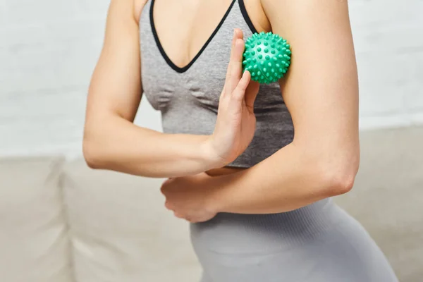 Cropped view of woman in sportswear massaging muscle on arm with manual massage ball in blurred living room, balancing energy and holistic healing concept, myofascial release — Stock Photo