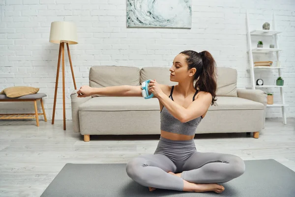 Young brunette woman in sportswear massaging arm with handled massager while sitting on fitness mat at home in living room, balancing energy and holistic healing concept, myofascial release — Stock Photo