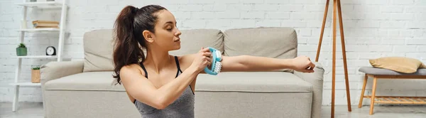Relaxed brunette woman in sportswear massaging muscle on arm with handled massager in blurred living room at home, home-based massage and holistic wellness practices concept, banner — Stock Photo