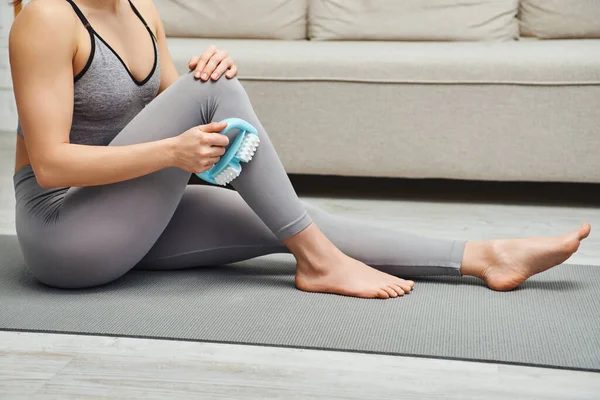 Cropped view of young woman in activewear using handled massager on leg while sitting on fitness mat near couch in living room at home, inner peace and harmony concept, myofascial release — Stock Photo