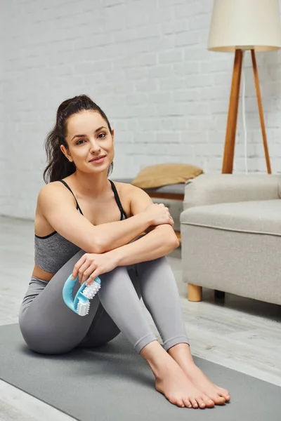Smiling and barefoot brunette woman in activewear holding handled massager and looking at camera while sitting on fitness mat in living room at home, inner peace and harmony concept — Stock Photo