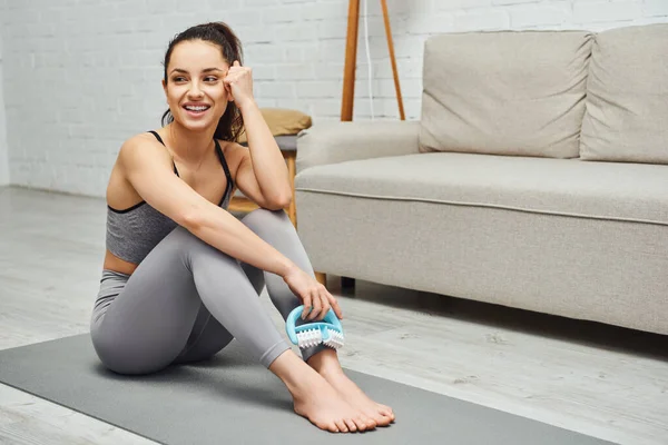 Cheerful young brunette woman in activewear holding handled massager and looking away while sitting on fitness mat on floor in living room at home, inner peace and harmony concept — Stock Photo