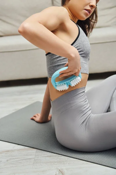 Cropped view of young brunette woman in activewear massaging body with handled massager and sitting on fitness mat at home, therapeutic tool for massage and holistic wellness practices concept — Stock Photo