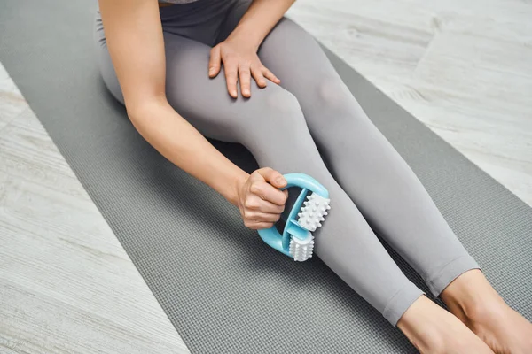 Cropped view of young woman in activewear massaging muscle on leg with handled massager and sitting on fitness mat at home, therapeutic tool for massage and holistic wellness practices concept — Stock Photo