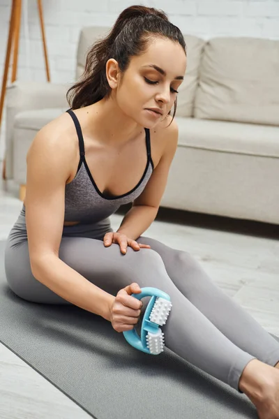 Young brunette woman in sportswear massaging muscle on leg with handled massager while sitting on fitness mat in living room, therapeutic tool for massage and holistic wellness practices concept — Stock Photo