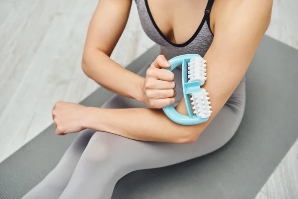 Draufsicht auf junge Frau in Sportbekleidung, die Muskeln am Arm mit Handmassagegerät massiert und zu Hause auf Fitnessmatte sitzt, Therapiewerkzeug für Massage und ganzheitliches Wellness-Praxiskonzept — Stockfoto