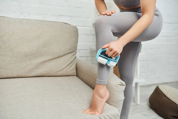 Cropped view of young barefoot woman in activewear massaging leg with handled massager doing lymphatic drainage near couch, therapeutic tool for massage and holistic wellness practices concept — Stock Photo