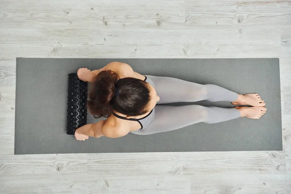 Vue aérienne de la jeune femme brune en vêtements de sport tenant un masseur à rouleaux assis sur un tapis de fitness à la maison, concept de massage à domicile et de pratiques holistiques, libération myofasciale — Photo de stock