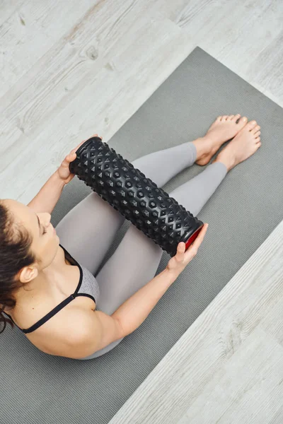 Overhead view of young barefoot woman in activewear holding roller massager and sitting on fitness mat in living room, home-based massage and holistic practices concept, myofascial release — Stock Photo