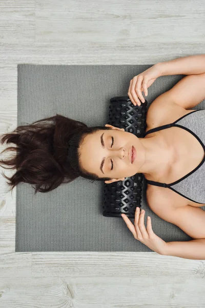Вид зверху на молоду розслаблену брюнетку в спортивному одязі, використовуючи роликовий масажер на шиї, лежачи на фітнес-матусі вдома, концепція домашнього масажу та цілісних практик, міофасциальний випуск — стокове фото