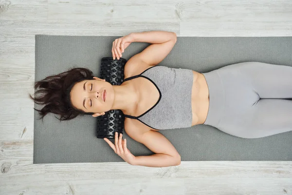 Top view of young brunette woman in sportswear using roller massager on neck while lying on fitness mat at home, home-based massage and holistic practices concept, myofascial release — Stock Photo