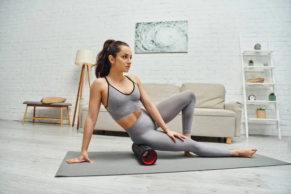 Young brunette and barefoot woman in sportswear looking away while massaging hip with roller massager on fitness mat in living room at background, focus on self-care and well-being concept — Stock Photo