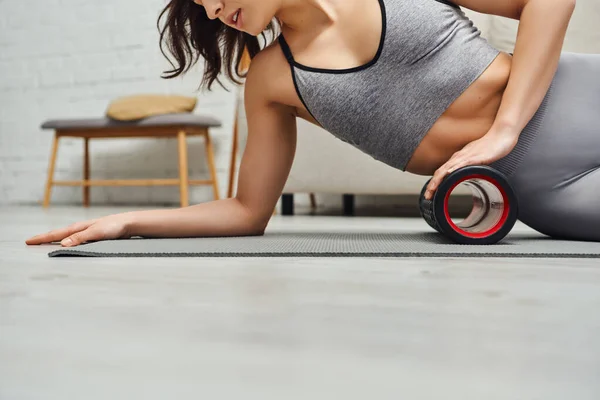 Cropped view of brunette woman in activewear massaging body with modern roller massager while lying on fitness mat at home, focus on self-care and well-being concept, tension relief — Stock Photo