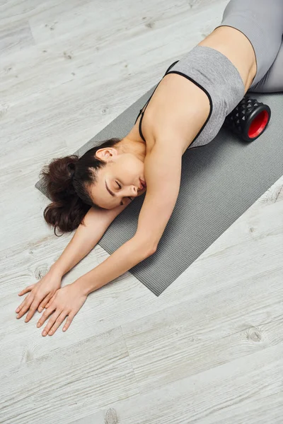 Top view of relaxed and peaceful young brunette woman massaging belly with roller massager and lying on fitness mat at home, focus on self-care and well-being concept, tension relief — Stock Photo