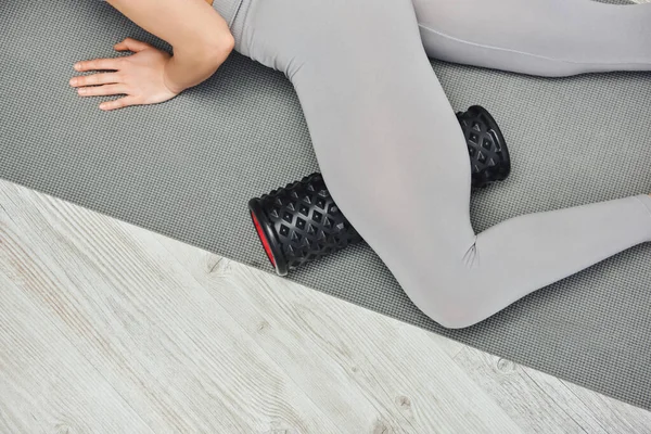 Cropped view of young woman in activewear massaging leg with roller massager and lying on fitness mat at home, focus on self-care and well-being concept, tension relief — Stock Photo