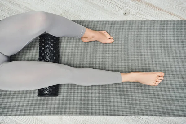 Cropped view of barefoot woman in sportswear massaging legs with modern roller massager and lying on fitness mat at home, maintaining healthy lymphatic system concept, tension relief — Stock Photo