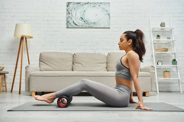 Side view of relaxed brunette woman in fitness clothes massaging legs with roller massager and sitting on mat in living room at home, maintaining healthy lymphatic system concept, myofascial release — Stock Photo