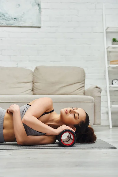 Entspannte junge Frau in Sportbekleidung schließt die Augen, während sie zu Hause den Nacken mit einem modernen Rollmassager auf der Fitnessmatte massiert, das Gefühl der Ruhe und fördert das Entspannungskonzept, die myofasziale Befreiung — Stockfoto