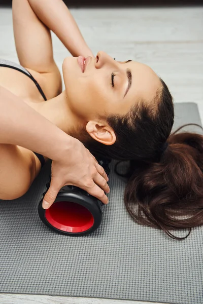 Ritratto di giovane donna rilassata e bruna che massaggia il collo per la circolazione linfatica con massaggiatore a rulli sul tappeto fitness a casa, senso di tranquillità e promuovere il concetto di relax — Foto stock