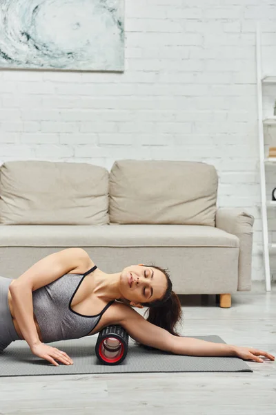 Mujer morena joven en ropa deportiva relajante mientras masajea axila con masajeador de rodillos en la alfombra de fitness en el suelo en la sala de estar en casa, sensación de tranquilidad y promover el concepto de relajación - foto de stock