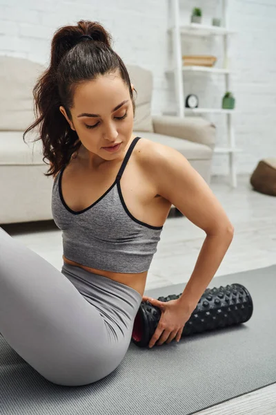 Young brunette woman in activewear holding modern roller massager for lymphatic drainage and sitting on fitness mat at home, sense of tranquility and promote relaxation concept, tension relief — Stock Photo