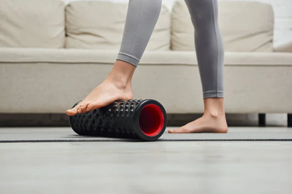 Cropped view of barefoot woman in sportswear massaging feet with roller massager and standing on fitness mat near couch at home, promoting lymph flow and wellness at home concept, tension relief — Stock Photo