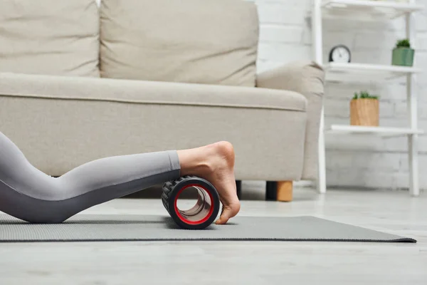 Cropped view of barefoot woman in activewear massaging legs with roller massager on fitness mat near couch in living room at home, promoting lymph flow and wellness at home concept, tension relief — Stock Photo