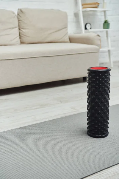 Modern black roller massager standing on fitness mat near blurred couch in living room at home, promoting lymph flow and wellness at home concept — Stock Photo