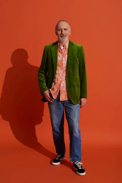 Full length of senior male model in green velour blazer posing with thumb in pocket of blue denim jeans on red orange background, smiling face, personal style, positive aging concept — Stock Photo