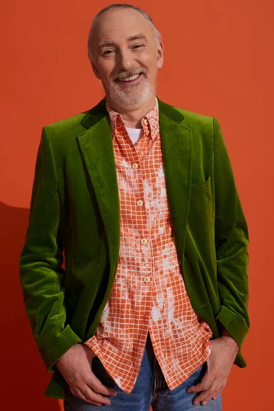Homem idoso despreocupado, cabelos grisalhos e elegante em blazer de veludo verde e camisa da moda de pé com polegares nos bolsos e sorrindo para a câmera no fundo laranja vermelho, conceito de envelhecimento feliz — Fotografia de Stock