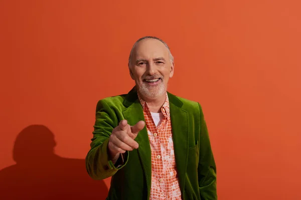 Счастливая старость, сияющая улыбка, веселый пожилой человек с седыми волосами и бородой, в модной рубашке и зеленом велюровом блейзере, смотрящем в камеру и указывающим пальцем на красный оранжевый фон — стоковое фото