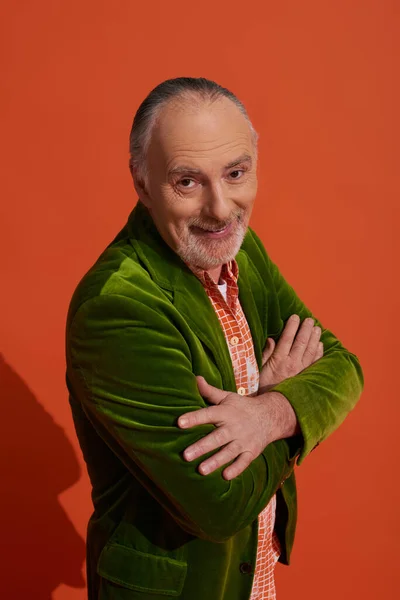 Optimistischer und charismatischer bärtiger Senior, der mit verschränkten Armen vor rot-orangefarbenem Hintergrund in die Kamera lächelt, modischer Look, persönlicher Stil, grüner Velours-Blazer, Happy-Aging-Konzept — Stockfoto