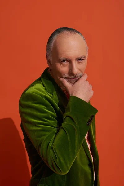 Hombre anciano reflexivo y positivo tocando la barba y mirando a la cámara en el fondo rojo anaranjado, ropa casual, chaqueta de terciopelo verde, mirada expresiva, concepto de estilo de vida de moda - foto de stock