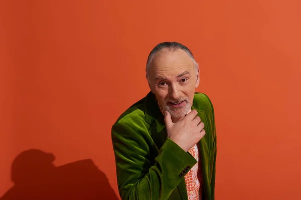 Fashionable and charismatic senior man in green velour blazer touching beard and smiling at camera while posing on red background, happy emotion, positive and trendy aging concept — Stock Photo