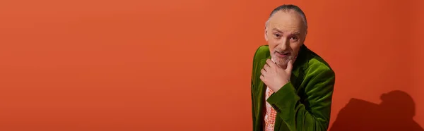 Homme âgé curieux et joyeux avec cheveux gris et barbe tenant la main près du menton et regardant la caméra sur fond rouge orange, modèle plus ancien, veste en velours vert, vieillissement positif et élégant, bannière — Photo de stock